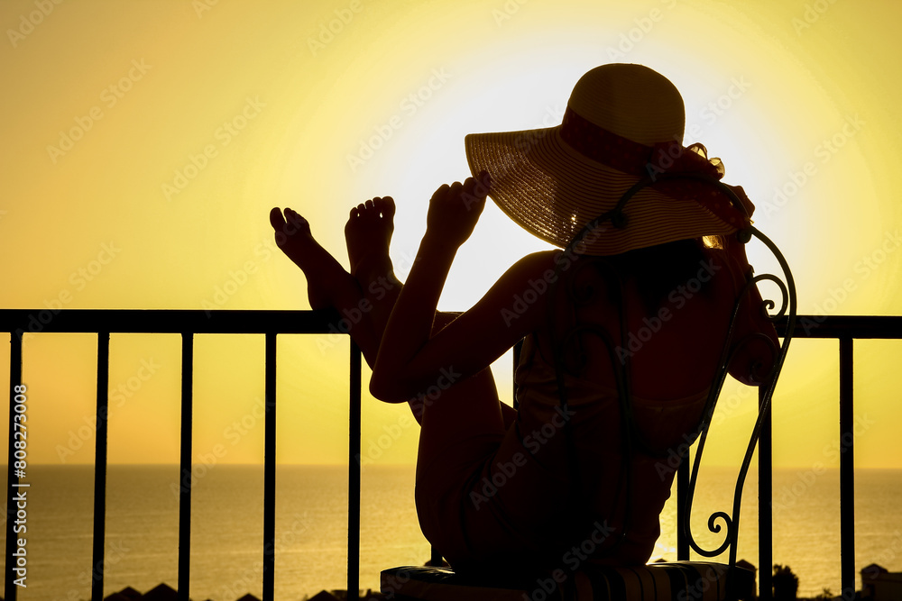 Silhouette of a girl on a chair in the loggia balcony background. A happy woman with a hat on vacation while traveling.