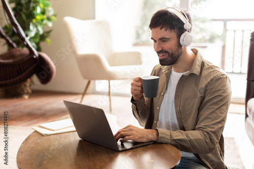 A man is seated at a table, focused on his laptop screen. Next to him is a cup of coffee, steam rising. The mans hands are typing on the keyboard as he works.