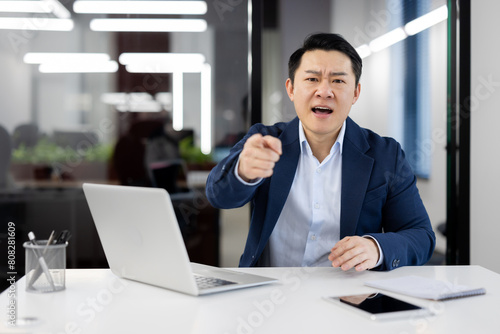 Angry young asian man and businessman, boss, director sitting in office at the table and unsatisfied shouting at pointing finger at camera.