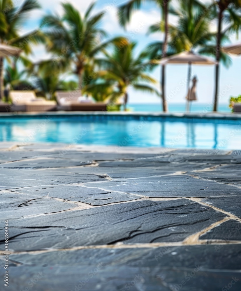 Close up of black stone floor edge near luxury outdoor swimming pool with palm trees and chairs on background