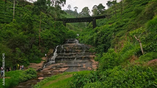 Drone of Nanu Oya viaduct over cascading waterfall amidst plants. Train crosses historic bridge, scenic travel route in Sri Lanka. Tourists visit, admire tea plantation landscape from viewpoint Aerial photo