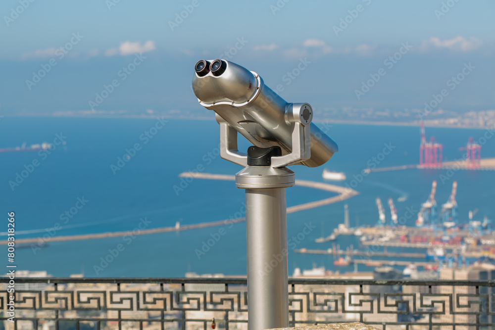 Coin operated binocular is mounted on a viewpoint, view of a bustling harbor, port and sea in Haifa, Israel. The intricate railing and blurred background highlight the binoculars as the main focus