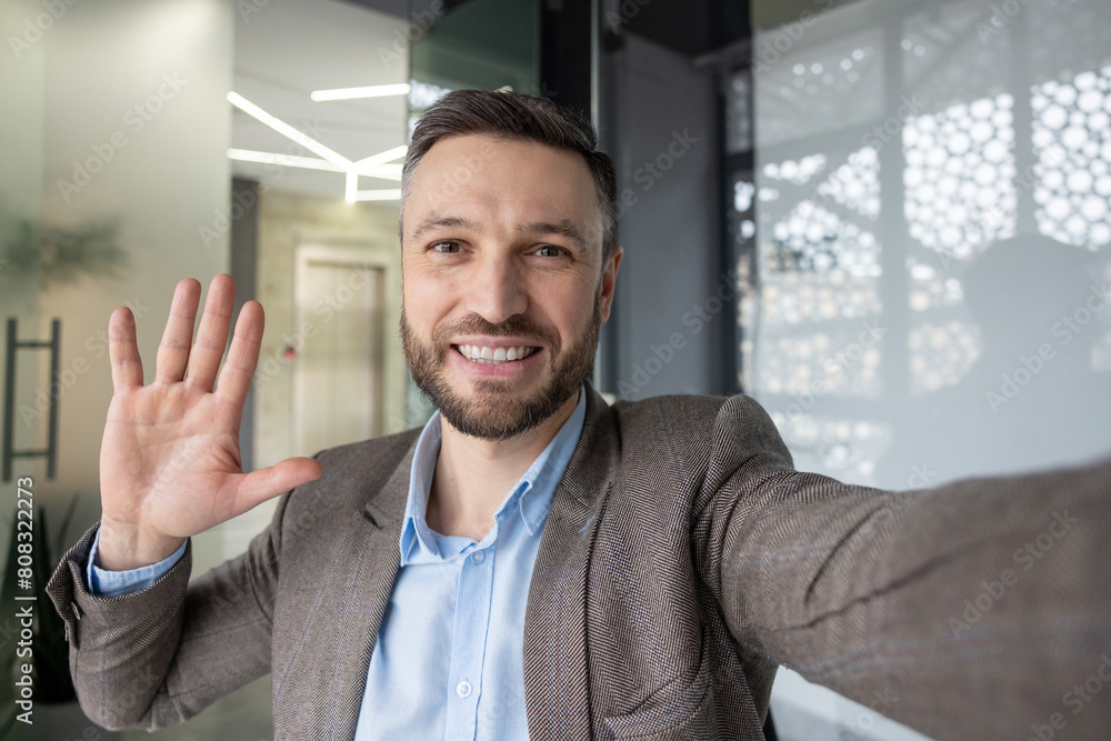 Cheerful professional man in a suit greeting someone via a video call. He waves while holding his phone, showcasing modern communication. Cheerful professional man in a suit greeting someone via a video call. He waves while holding his phone, showcasing modern communication.