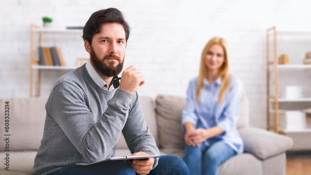 A bearded man therapist, holding a notebook, is seated attentively with a contemplative expression, while a lady client sits in the background on a couch A bearded man therapist, holding a notebook, is seated attentively with a contemplative expression, while a lady client sits in the background on a couch