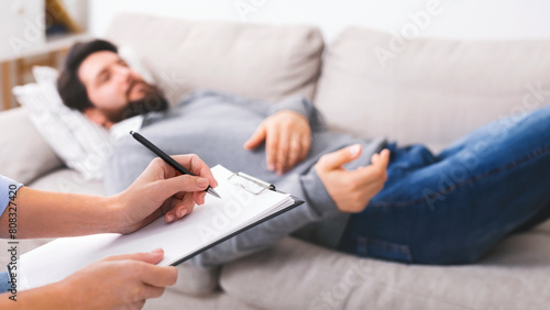 Stressed man telling about his problems to doctor, lying on couch at psychotherapist office, doctor making notes photo