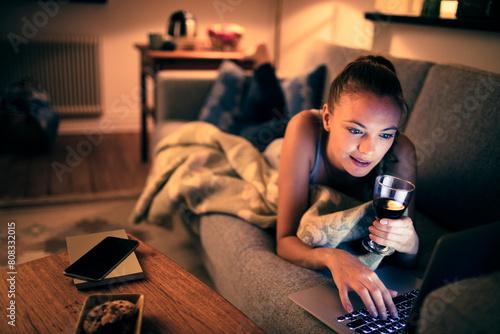 Relaxed young woman with wine using laptop at home in the evening photo