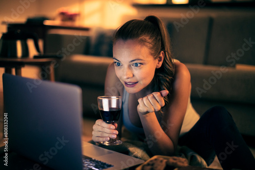 Relaxed young woman with wine using laptop at home in the evening photo