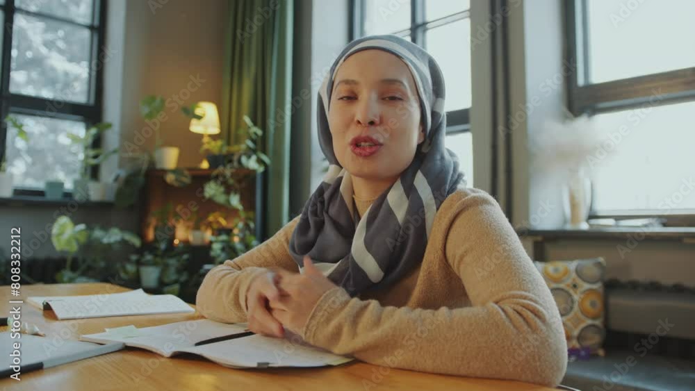 Young Muslim woman in hijab sitting at desk with copybooks and speaking on camera during e-meeting or online lesson via video call