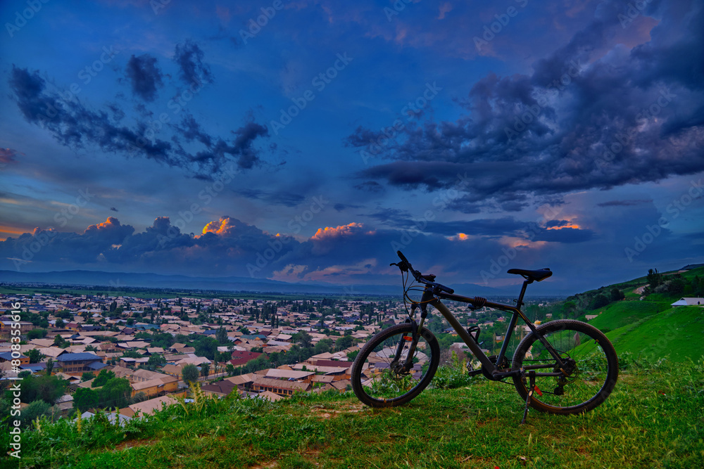 Obraz premium A mountain bike standing atop a green hill with a city panorama against a stormy sky in the background. Jalal-Abad, Kyrgyzstan