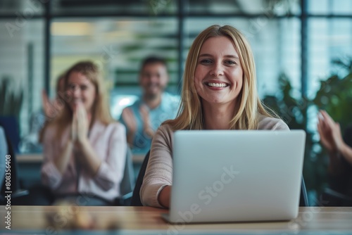 Cheerful young businesswoman works on her laptop, with blurry colleagues applauding behind her, celebrating success in a modern office setting