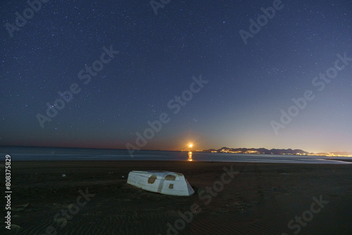 Sandy sea beach with white boat and illuminated Amalfi Coast at night with Orion and Pleiades constellation and moon rising, Paestum, Campania, Italy