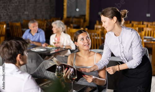Polite smiling waitress talking to positive young couple visiting restaurant for romantic dinner, recommending dishes from menu card