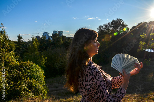 Woman wearing a dress with leopard print design holding fan of hundred dollars cash standing outdoors with greenery and buildings in the background illuminated by the setting sun. Financial success