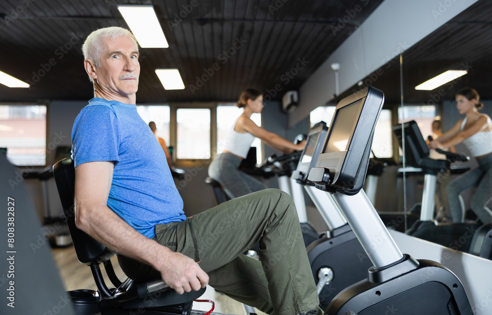 Elderly man, along with other people, is engaged on an stationary bike in gym