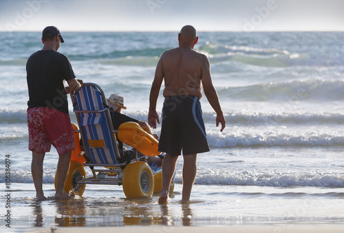 Two men are assisting a elderly man in a specialized beach wheelchair across the sandy shores. Sons taking care of their elderly parent with disabilities, spending time with them by the sea. Care