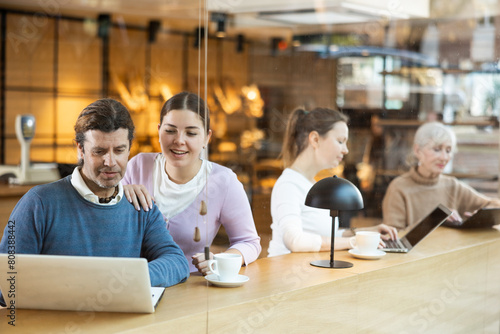 Cheerful young girl and adult man, friendly colleagues, discussing team project while sitting at table in cafe with laptop and cup of coffee