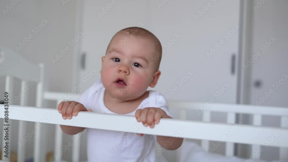 The baby is learning to stand. baby boy standing in the crib and holding on to the sides. Portrait of a newborn baby standing on his feet in a crib. Funny baby face. The baby is learning to stand. baby boy standing in the crib and holding on to the sides. Portrait of a newborn baby standing on his feet in a crib. Funny baby face.