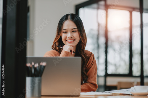 Contemplative young woman with a pen to her chin  deeply focused while working on her laptop in a sunlit office.