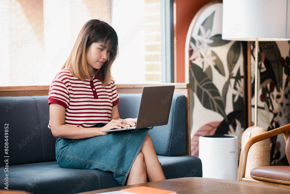 Fototapeta premium Concentrated young woman in a striped shirt working on a laptop while seated on a couch in a stylish cafe.