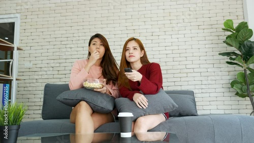 Two Women watch tv eat popcorn in living room together happy smiling face frienship relationship. woman sitting couch sofa use remote control turn on television enjoy watch movie home entertainment photo