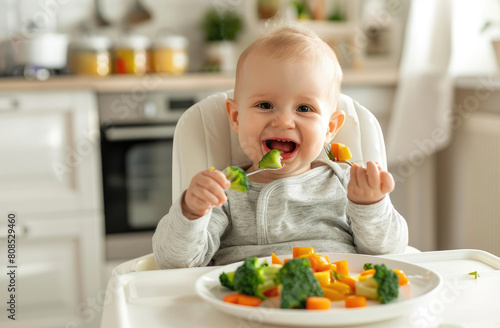 Cute baby sitting in a high chair and eating food, with a white kitchen background. The baby is smiling while holding his hand up to the camera.