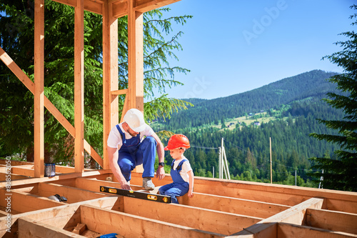 Father with toddler son building wooden frame house. Male builder demonstrating to his son how to measure level using spirit level on construction site on sunny day. Carpentry and family concept. photo