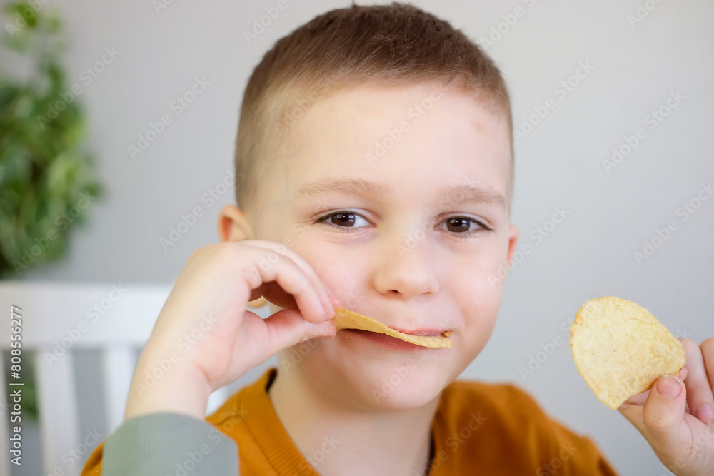Close-up of hungry child putting potato chips into his mouth while sitting at table. Boy happily eats fast food, junk food at home. Childhood obesity concept. Selective focus. Close-up of hungry child putting potato chips into his mouth while sitting at table. Boy happily eats fast food, junk food at home. Childhood obesity concept. Selective focus.