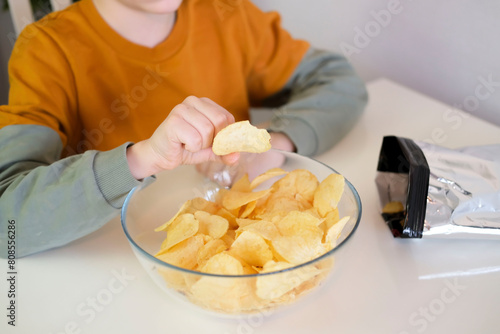 Close-up of hungry child putting potato chips into his mouth while sitting at table. Boy happily eats fast food, junk food at home. Childhood obesity concept. Selective focus.