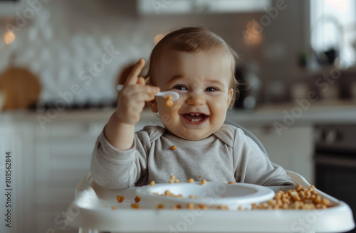 Cute baby sitting in a high chair and eating food  with a white kitchen background. The baby is smiling while holding his hand up to the camera.