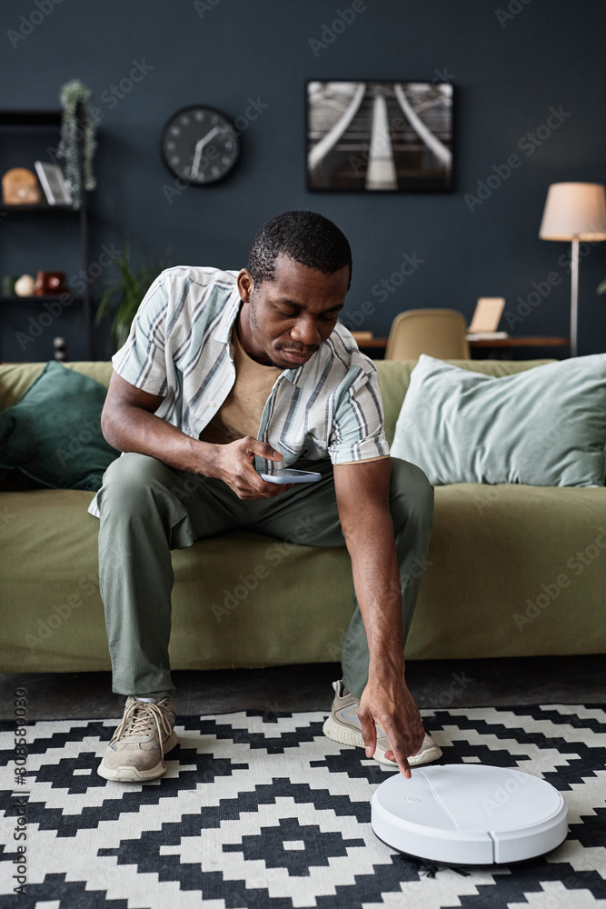 Fototapeta premium Vertical shot of young Black man sitting on sofa turning on modern robotic vacuum cleaner