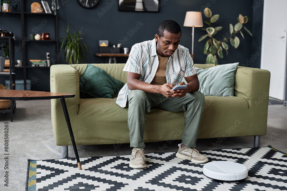 Fototapeta premium Long shot of African American man sitting on couch in modern living room adjusting robotic vacuum cleaner settings using app on smartphone