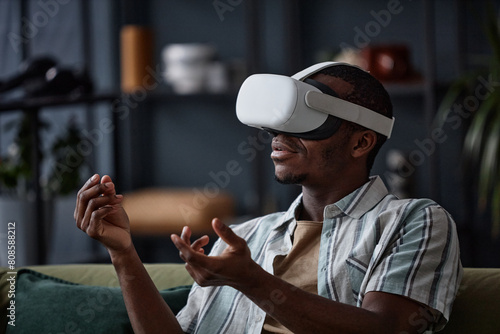 Medium closeup of young African American guy wearing VR headset sitting in living room playing video game, copy space