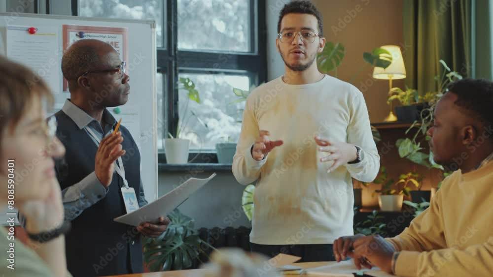 Young multiracial man standing by whiteboard and speaking in front of Black teacher and group mates during class in school for immigrant student Young multiracial man standing by whiteboard and speaking in front of Black teacher and group mates during class in school for immigrant student