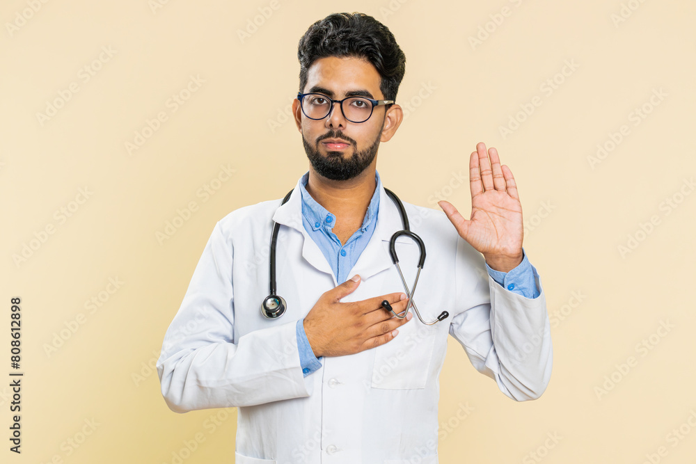 I swear to be honest. Sincere responsible Indian young doctor cardiologist man raising hand to take oath, promising to tell truth, keeping hand on chest. Apothecary pharmacy guy on beige background I swear to be honest. Sincere responsible Indian young doctor cardiologist man raising hand to take oath, promising to tell truth, keeping hand on chest. Apothecary pharmacy guy on beige background
