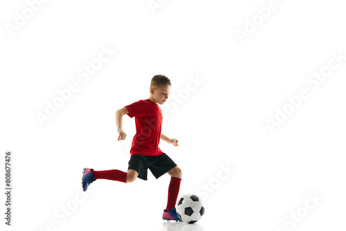 Little athlete boy, dribbling ball to take perfect goal against white studio background. Young football player in motion. Concept of professional sport, championship, youth league, hobby. Ad