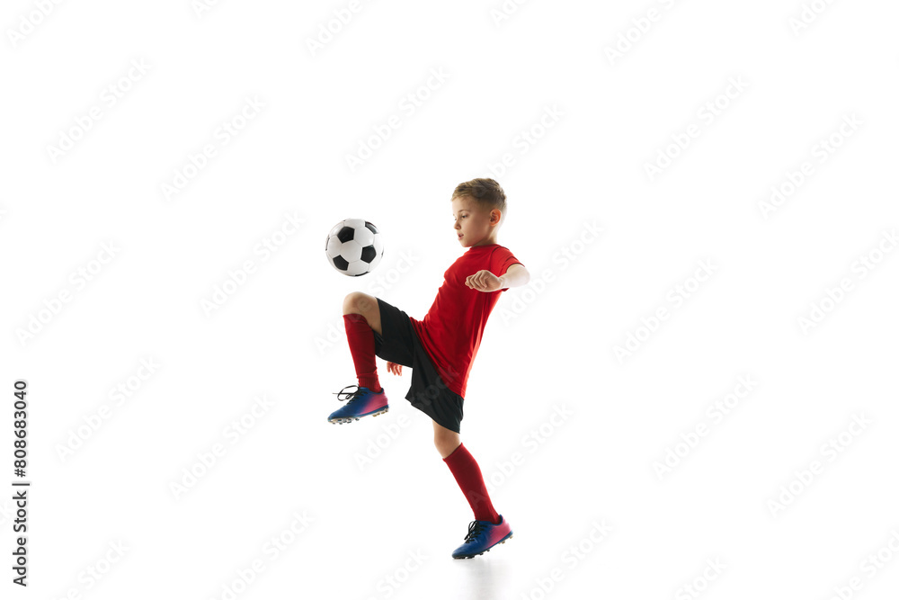 Dynamic portrait of little sporty boy with soccer ball kicking ball with knee in motion against white studio background. Concept of professional sport, championship, youth league, hobby. Ad