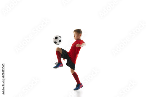 Dynamic portrait of little sporty boy with soccer ball kicking ball with knee in motion against white studio background. Concept of professional sport, championship, youth league, hobby. Ad © Lustre