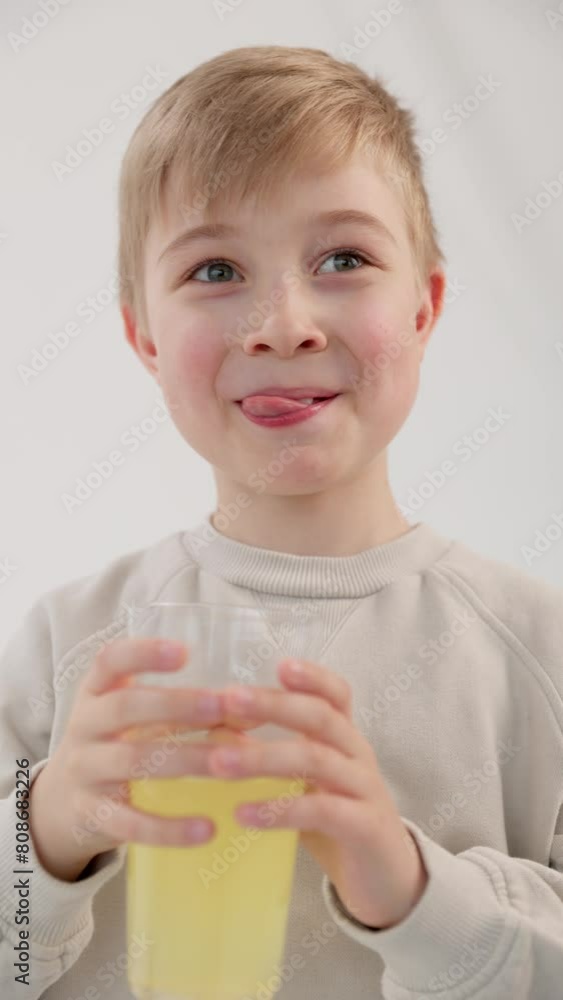 Vertical video. A blond Caucasian boy with blue eyes with a glass of lemonade smiles and licks his lips from the delicious drink. White background behind. The concept of healthy natural food. Vertical video. A blond Caucasian boy with blue eyes with a glass of lemonade smiles and licks his lips from the delicious drink. White background behind. The concept of healthy natural food.