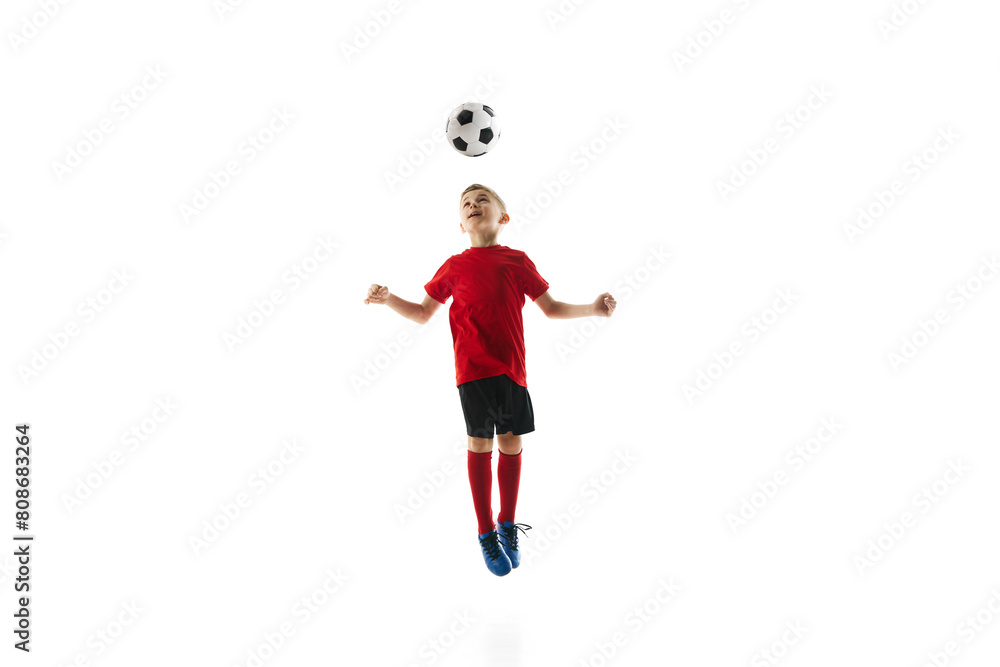 Dynamic image of young boy in mid-air, dressed in red soccer jersey and black shorts, skillfully heading soccer ball on white background. Concept of professional sport, championship, youth league. Ad