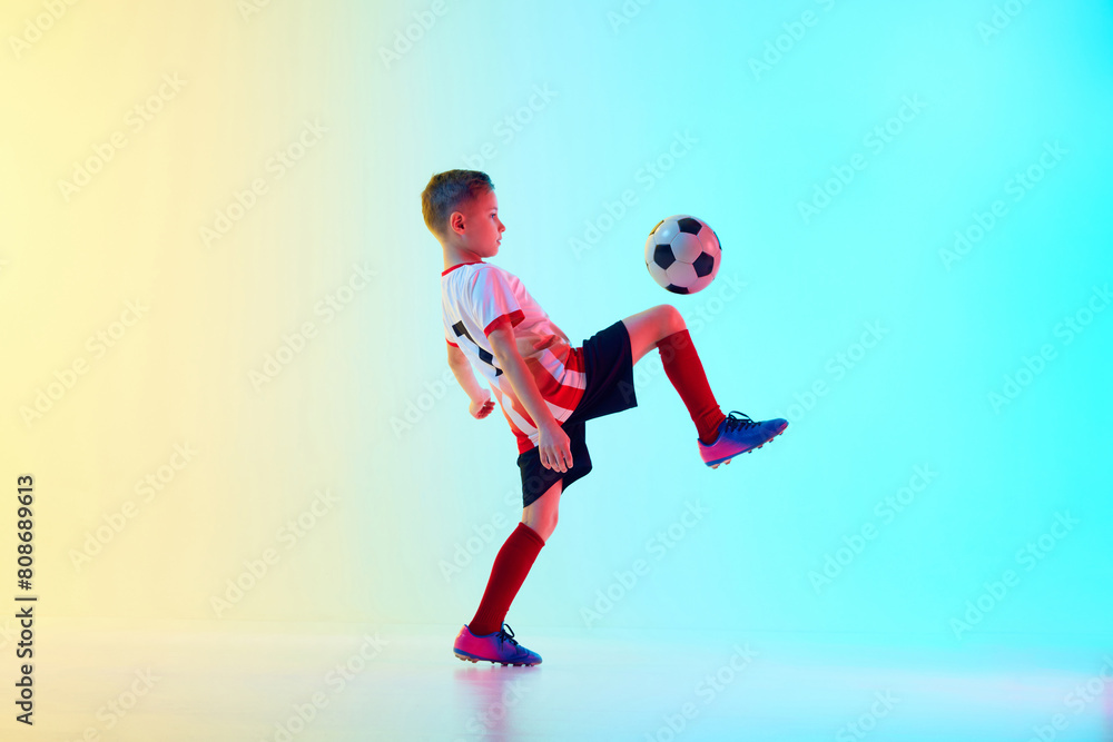 Dynamic photo of little athletic boy, soccer player kicking ball with knee in motion in neon light against gradient background. Concept of professional sport, championship, youth league, hobby. Ad