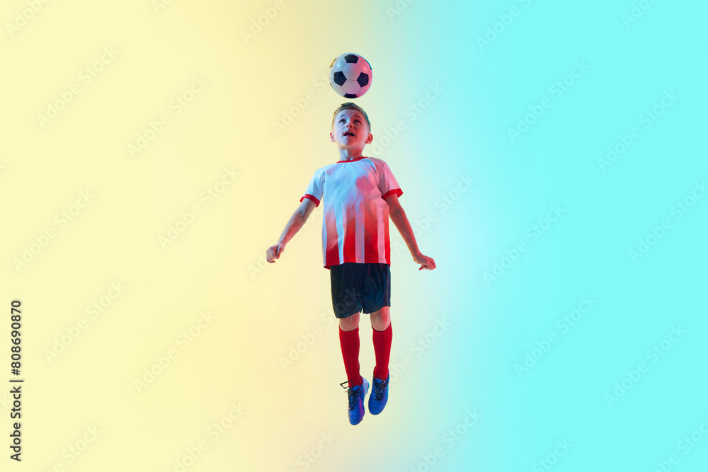 Dynamic image of young boy in mid-air, dressed in red soccer jersey, skillfully heading soccer ball in neon light against gradient background. Concept of professional sport, championship, youth league Dynamic image of young boy in mid-air, dressed in red soccer jersey, skillfully heading soccer ball in neon light against gradient background. Concept of professional sport, championship, youth league