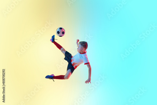 Dynamic photo of junior football player kicking ball in neon light against gradient background. Mid-air pass. Concept of professional sport, championship, youth league, hobby. Ad