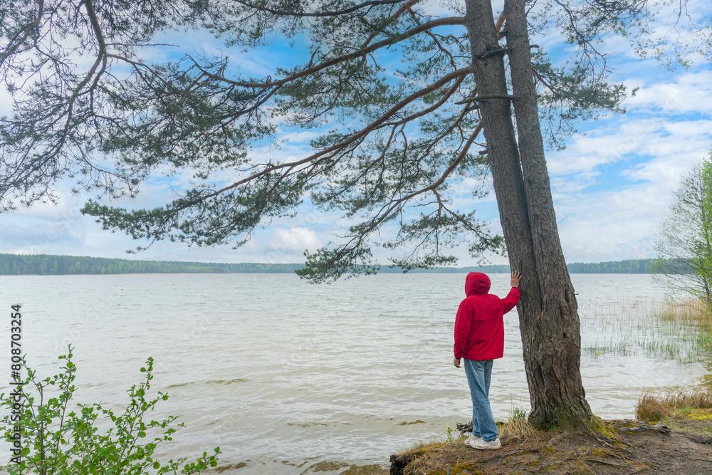 A tourist in a red windbreaker jacket stands on the shore of the lake.