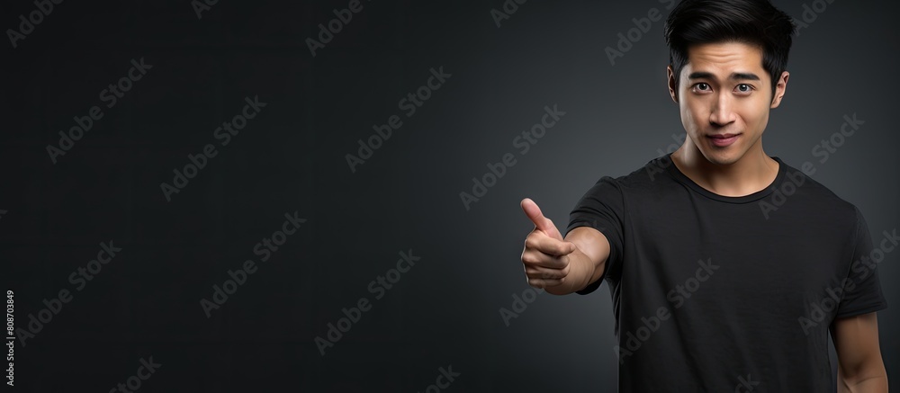 A young Asian man wearing a black t shirt looks directly at the camera with a funny expression while pointing towards the left side He is standing alone against a gray background Copy space image A young Asian man wearing a black t shirt looks directly at the camera with a funny expression while pointing towards the left side He is standing alone against a gray background Copy space image