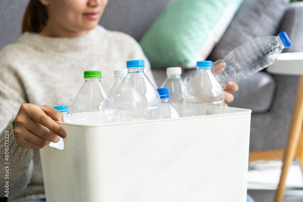 Fototapeta premium Happy Asian woman wearing a cream-colored sweater collects and sorts recycled plastic bottles into trash bins in the living room.