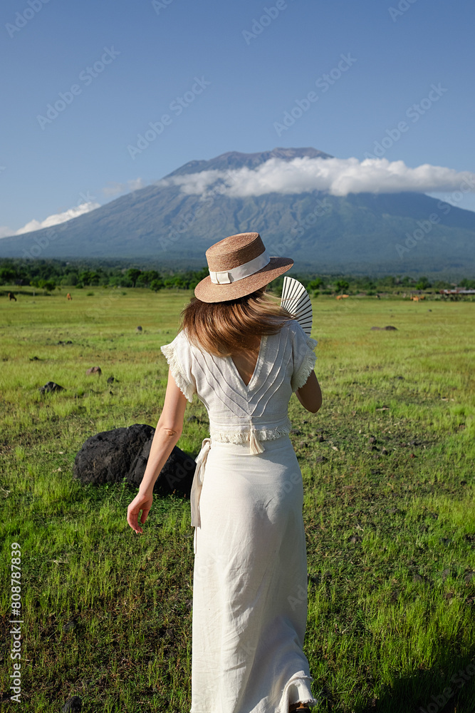 A young girl, wearing a hat and a white dress, stands on a green meadow with cows and admires the view of the mountain - Agung Volcano.