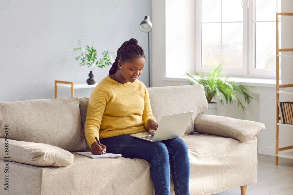 Happy woman enjoying online education in comfort of her own home. Young African American student girl sitting on sofa, using laptop computer, studying new useful information, and taking notes Happy woman enjoying online education in comfort of her own home. Young African American student girl sitting on sofa, using laptop computer, studying new useful information, and taking notes