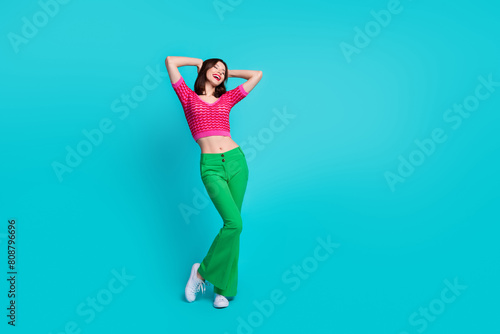 Full size photo of adorable gorgeous girl wear pink knit top green trousers hold arms behind head isolated on blue color background photo
