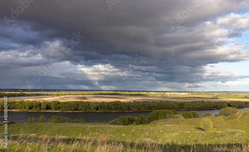  moment in a rural landscape  featuring a wide river meandering through fields and pastures. The horizon is dominated by a dramatic sky filled with storm clouds