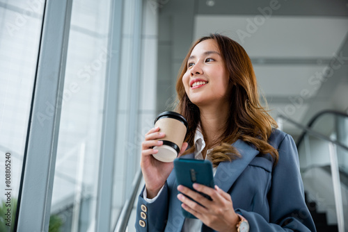 Young Asian businesswoman wearing blue shirt holding coffee cup and her smartphone. She smiling and looking out of the window and elevator blur background in the office. © sorapop